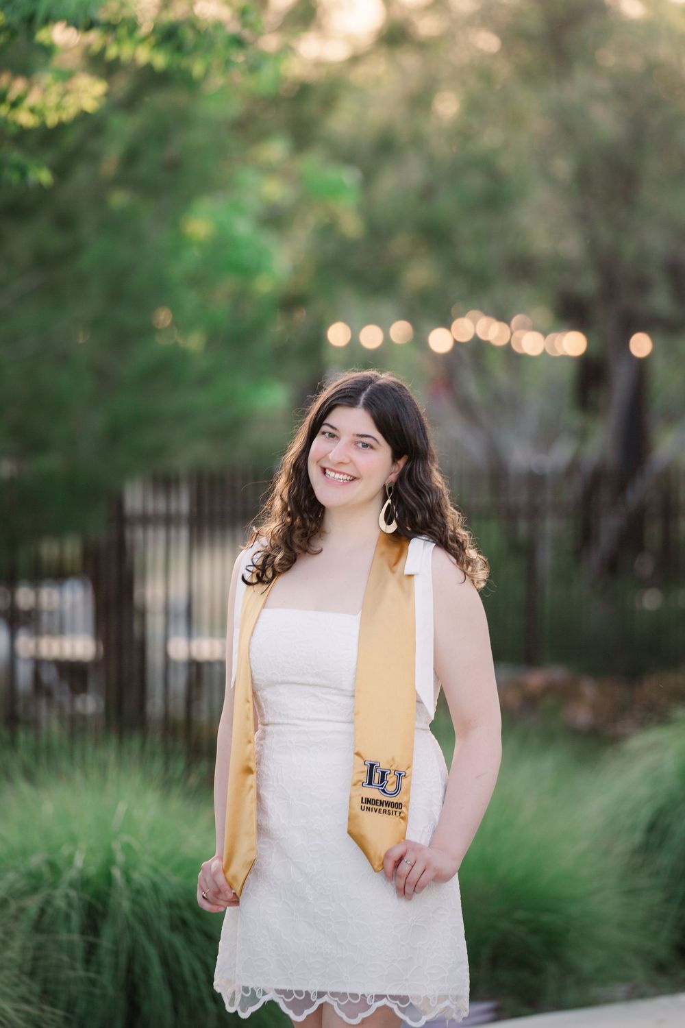 Graduate celebrates in white dress and gold stole outdoors with twinkling lights in background.