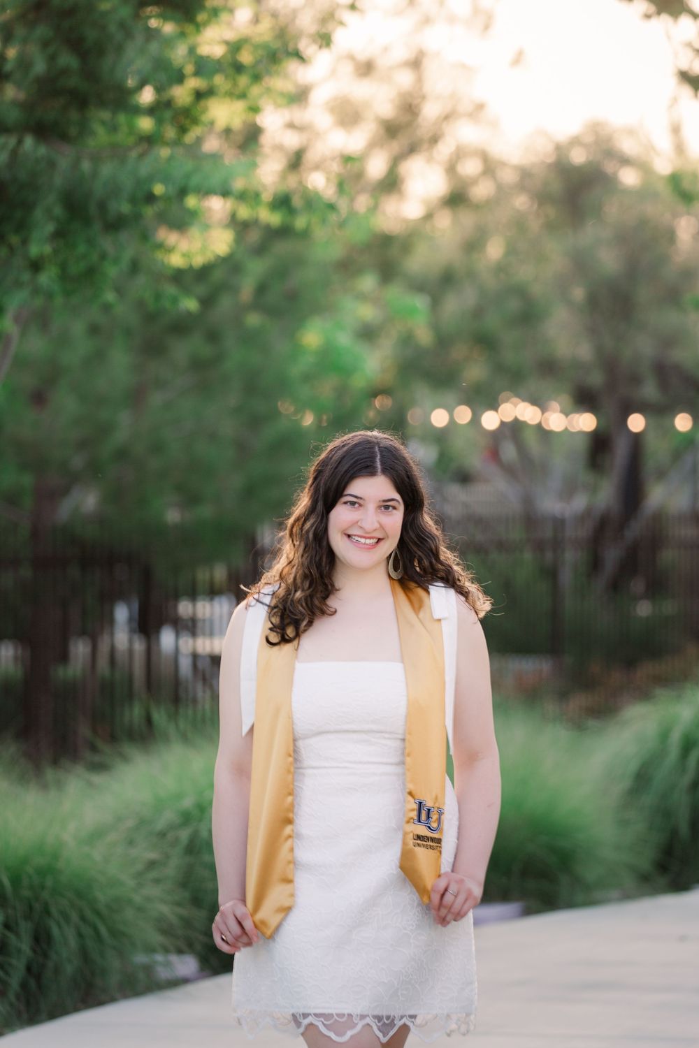 Graduate in white dress and gold stole smiles while walking through garden pathway.