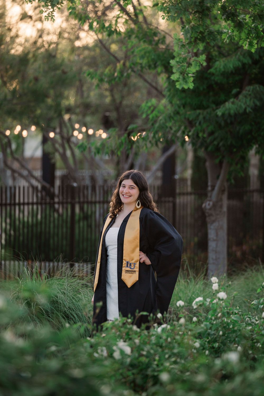 Graduate in academic regalia poses for photos in garden setting with string lights and trees in background.