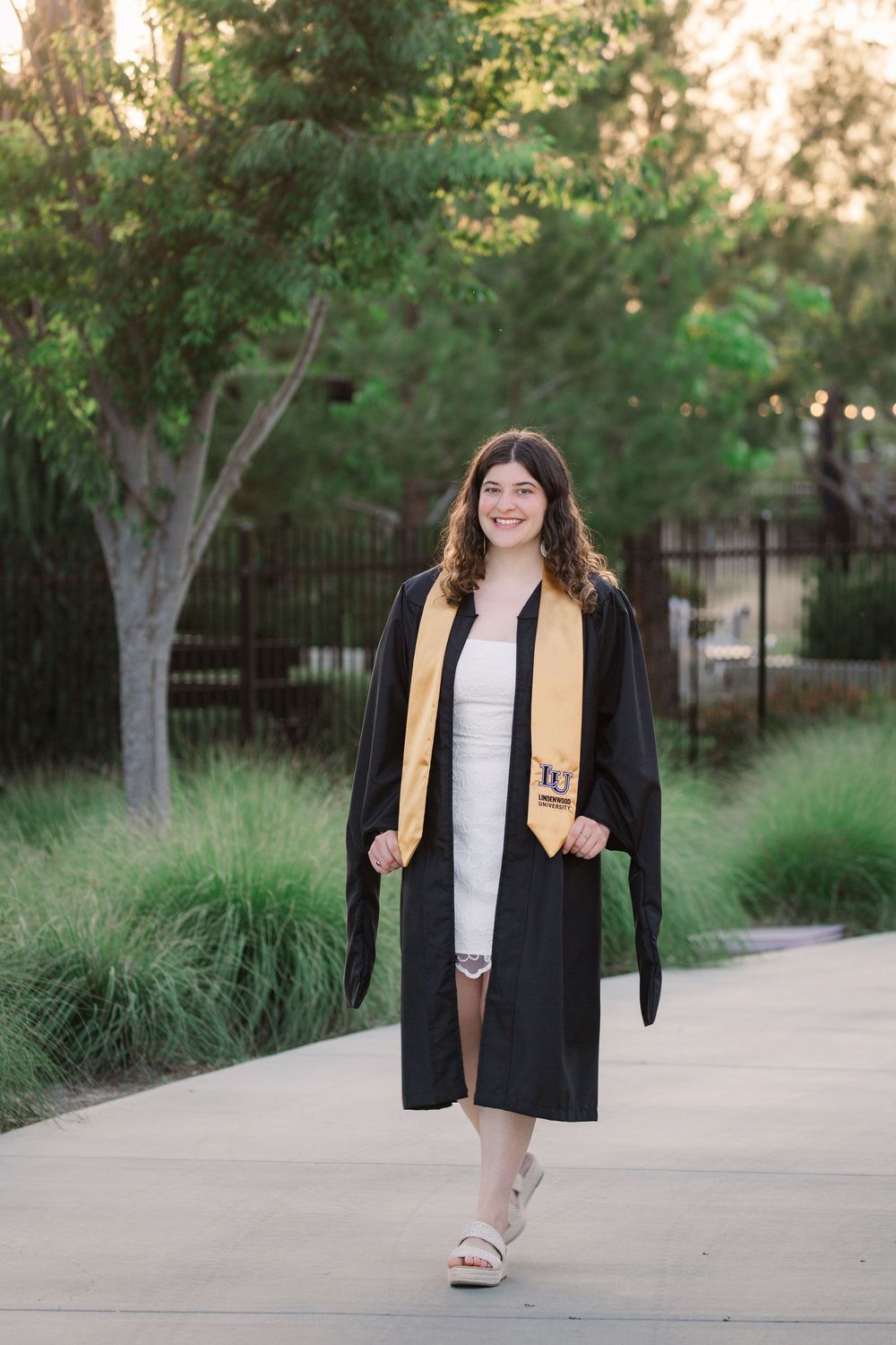 Graduate in black cap and gown with gold stole walks along outdoor pathway surrounded by greenery.