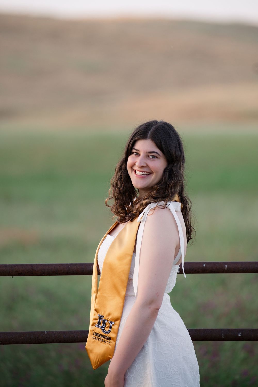 Person in a white sleeveless top and yellow backpack smiles while leaning against a rustic fence in an outdoor setting.
