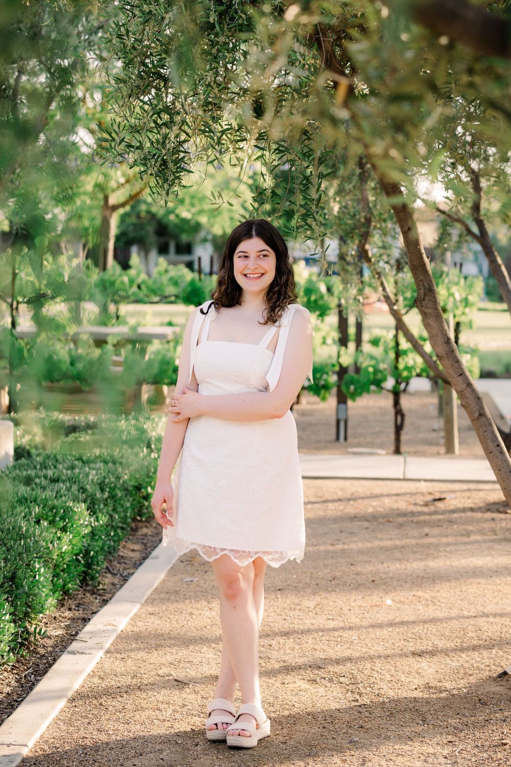 A person in a pink dress poses in a sunny park setting with trees and a dirt path.
