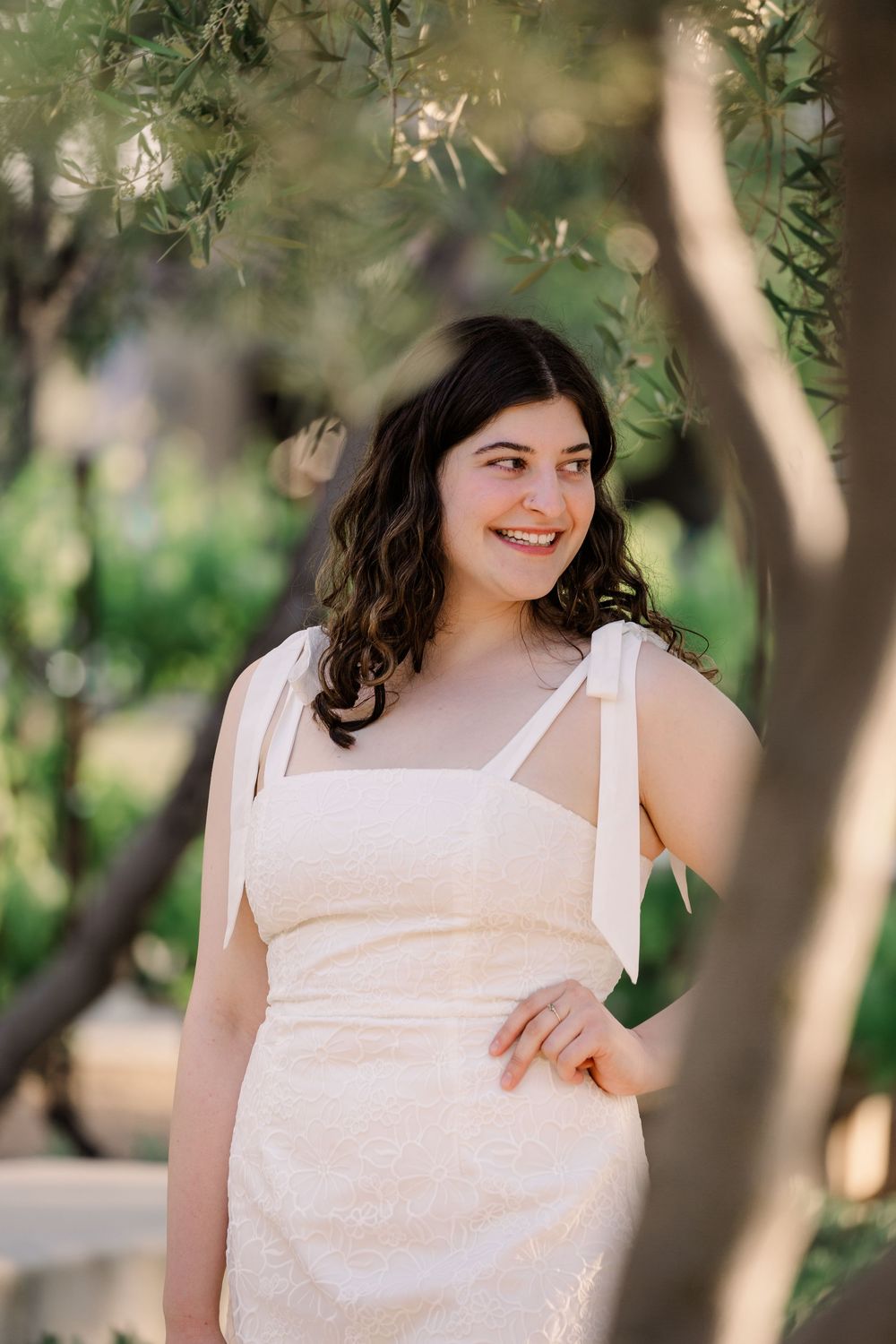Graduate in a white sundress poses outdoors under olive trees with natural bokeh lighting.