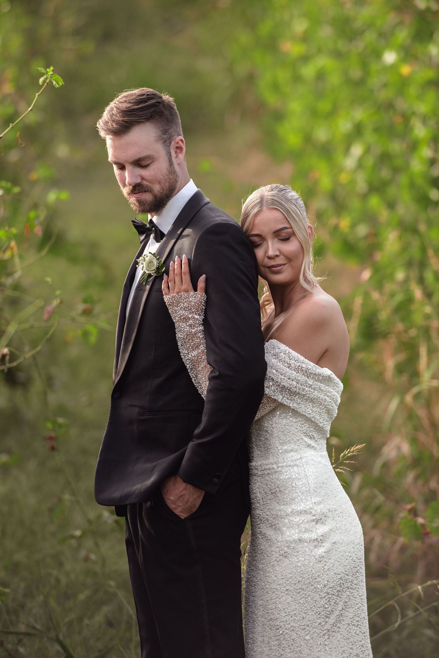 A bride in an off-shoulder white gown and groom in black tuxedo pose together outdoors among greenery.