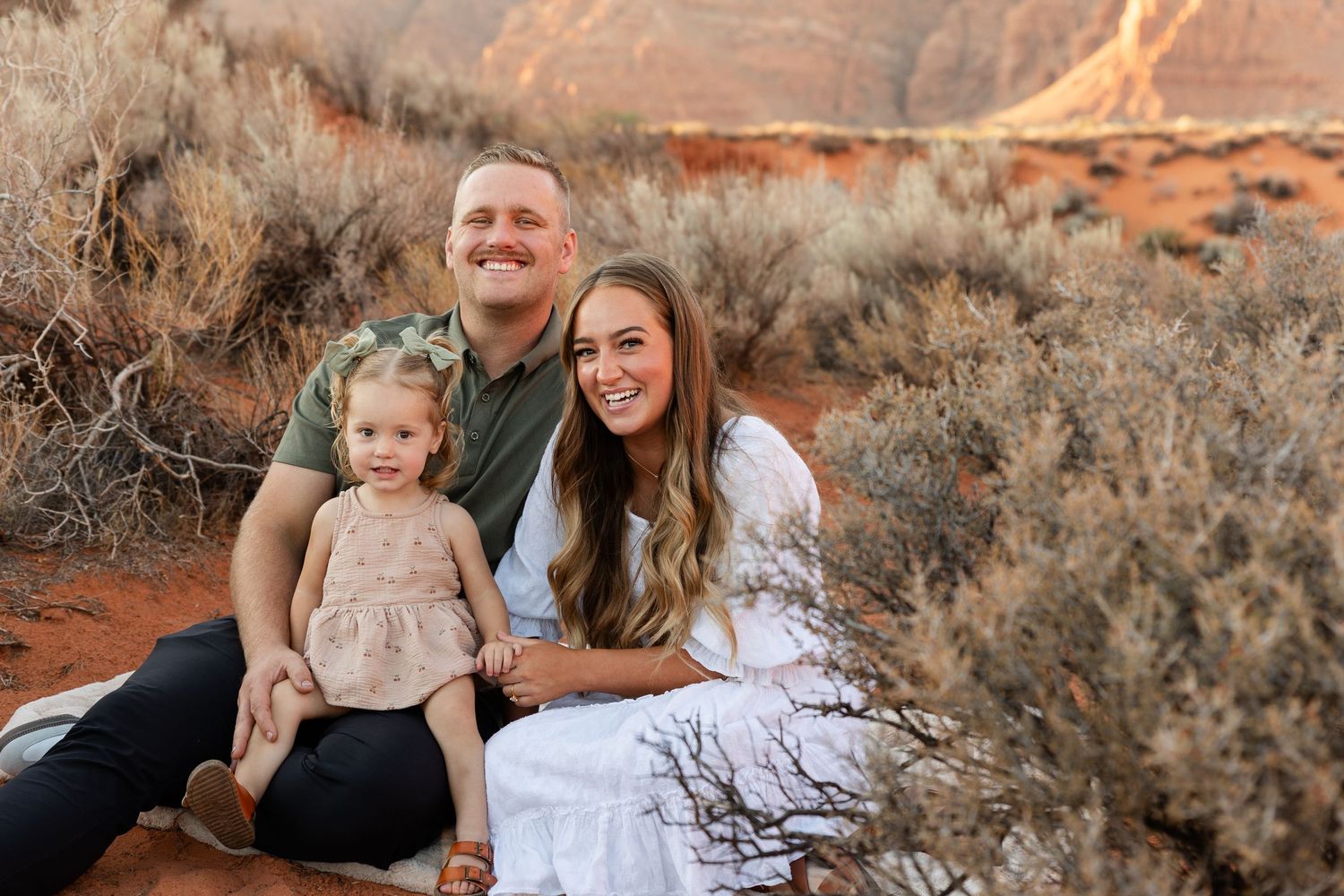 St. George Utah family photos of mom and dad sitting on a blanket with their 2-year-old daughter smiling in golden light with red rock back