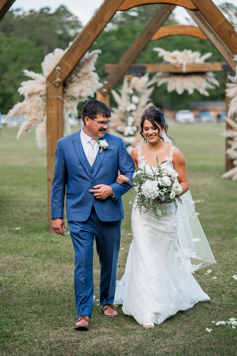 Couple walks together under a wooden arch in a garden setting with white floral decorations.