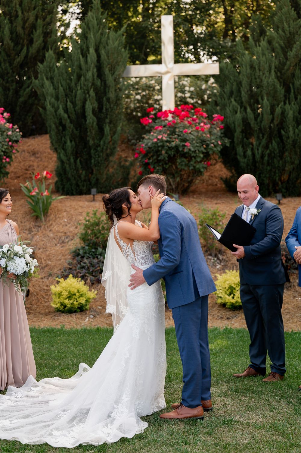 Wedding ceremony takes place under wooden arch with pink flowers and greenery in background.