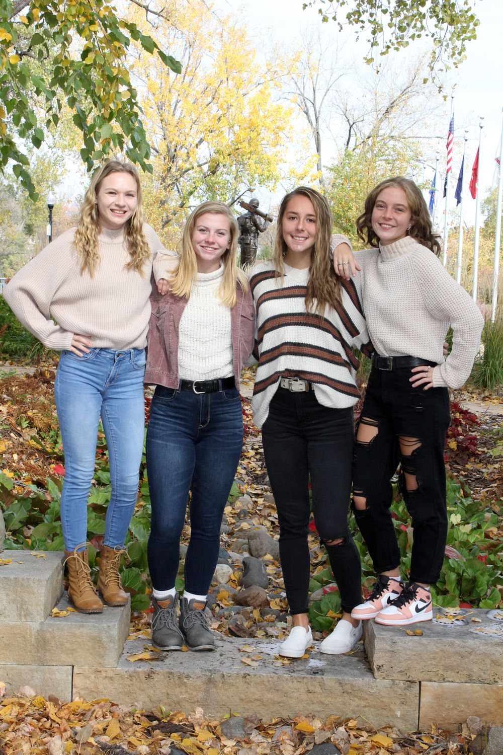 Four young women pose together outdoors in fall, wearing cozy sweaters and jeans, surrounded by autumn leaves and flags.