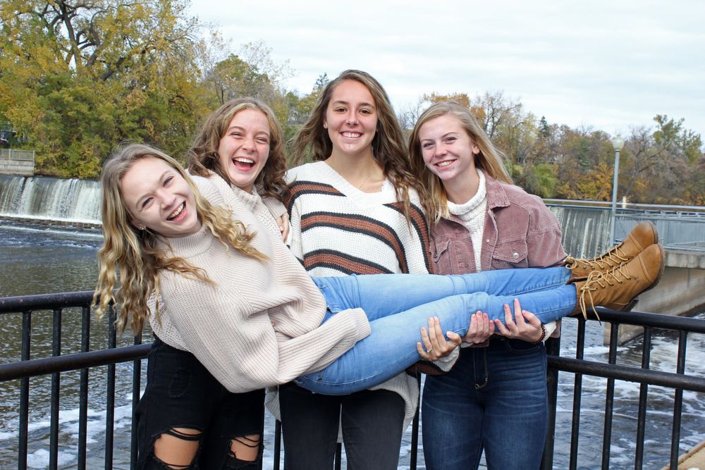 Four teenage girls laughing and posing outdoors near a waterfall, with one girl being held up by her three smiling friends.