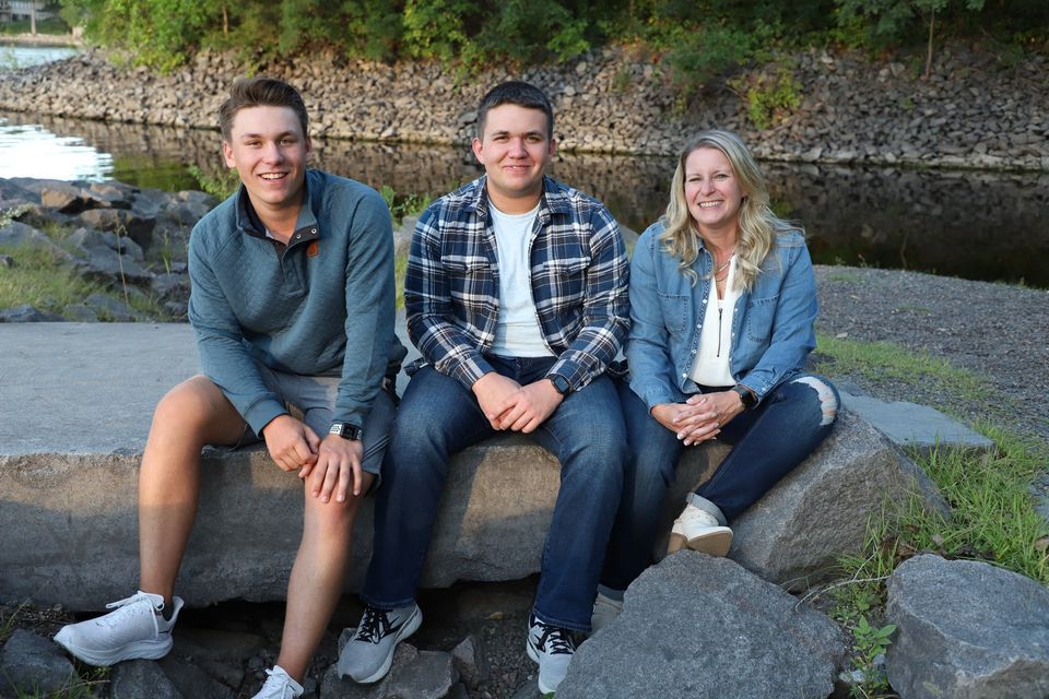 Three smiling friends sit on rocks near a river, wearing casual denim and light jackets in an outdoor setting.
