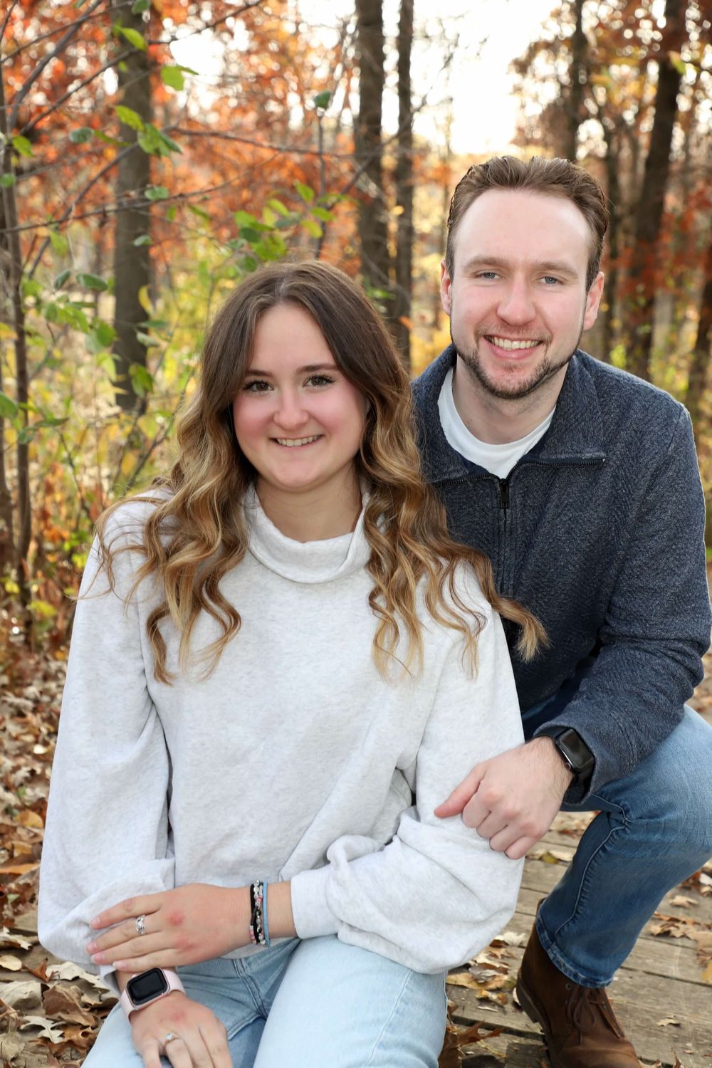 A smiling couple poses outdoors in autumn woods, woman in white turtleneck sweater and man in gray jacket.