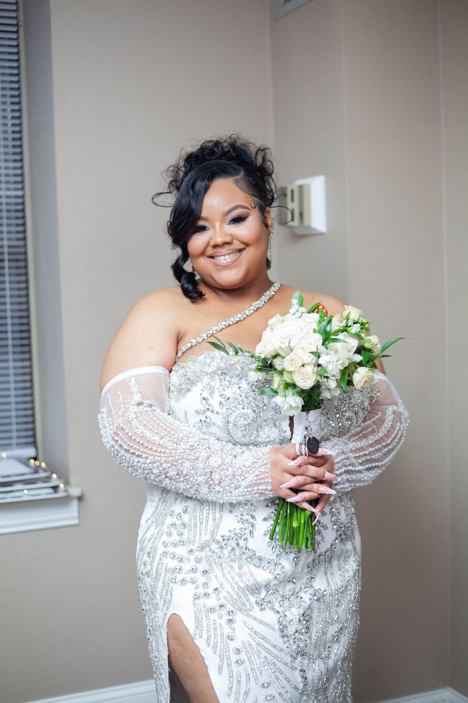 A bride in a beaded off-shoulder white wedding gown holds a white floral bouquet while posing in a room.
