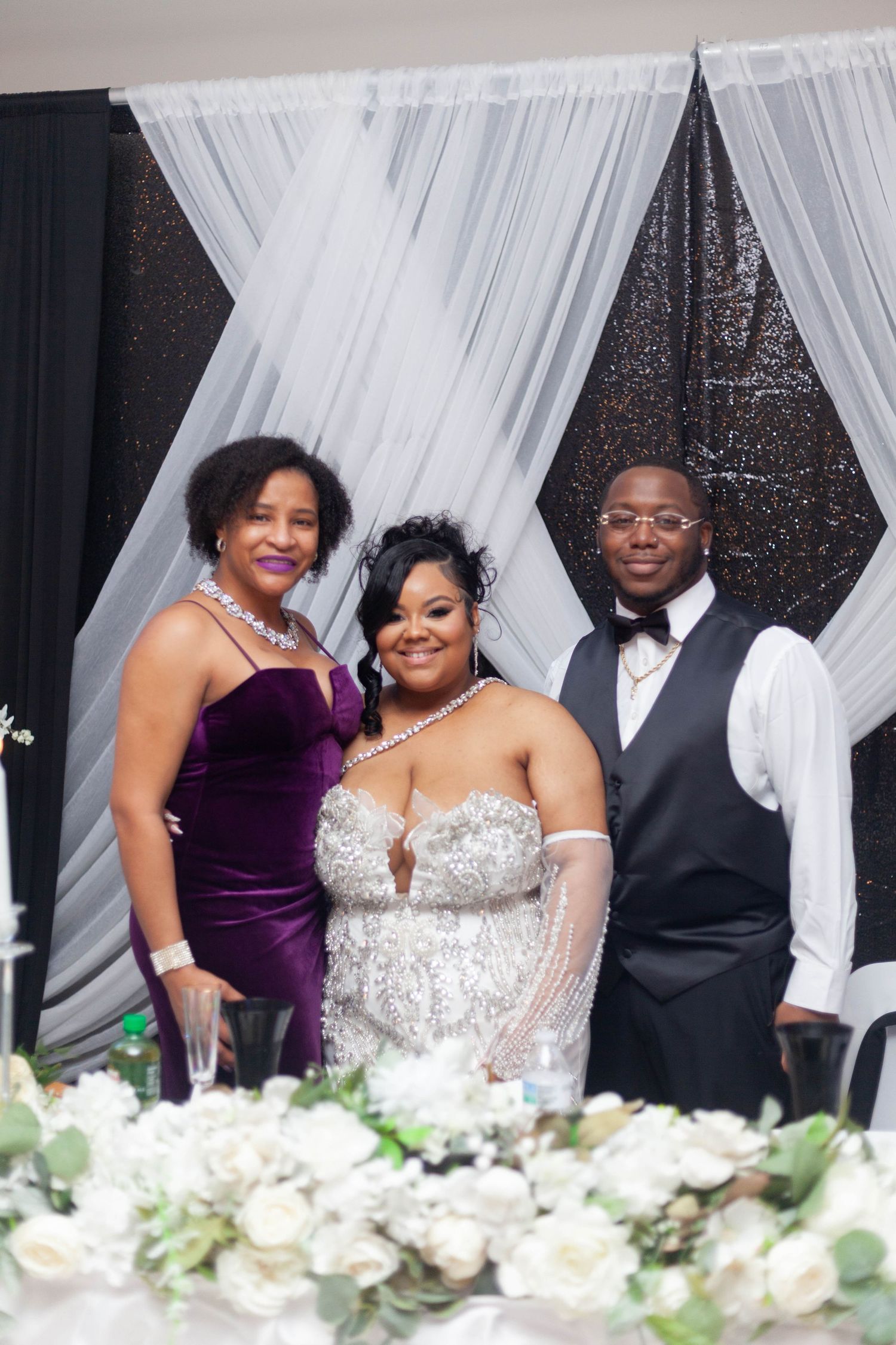 Wedding party members in purple dresses and formal attire pose together at an elegant reception with white drapes.