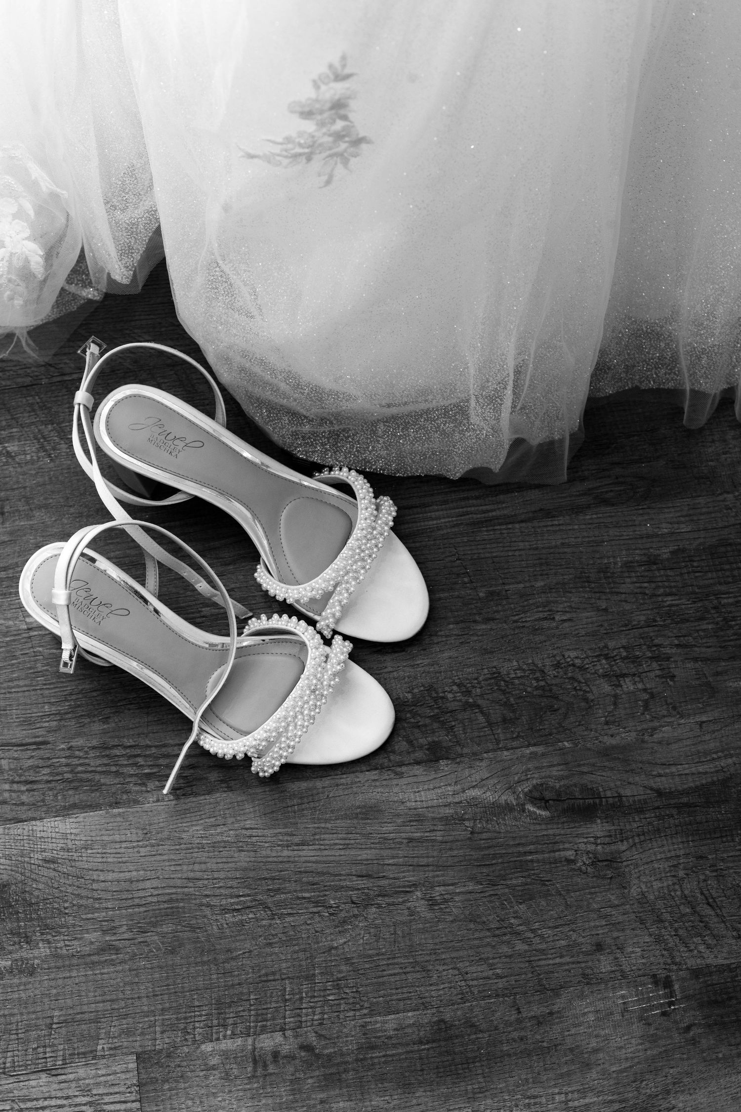 Black and white photo of elegant pearl-embellished bridal heels on wood floor beside a flowing white wedding dress.