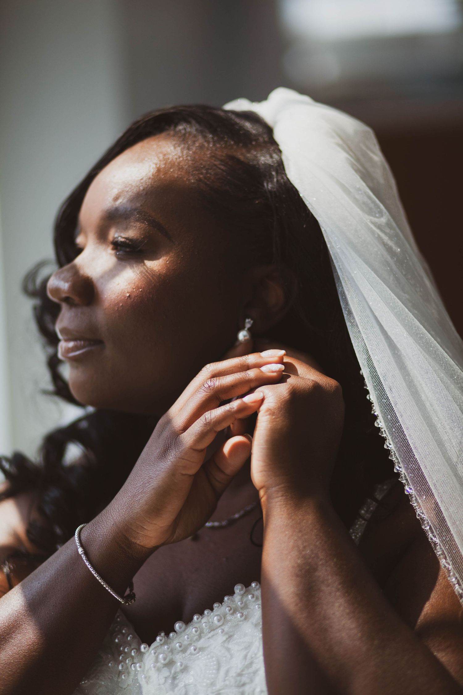 A bride in a white veil and lace gown gets her pearl earring fastened while preparing for her wedding day.