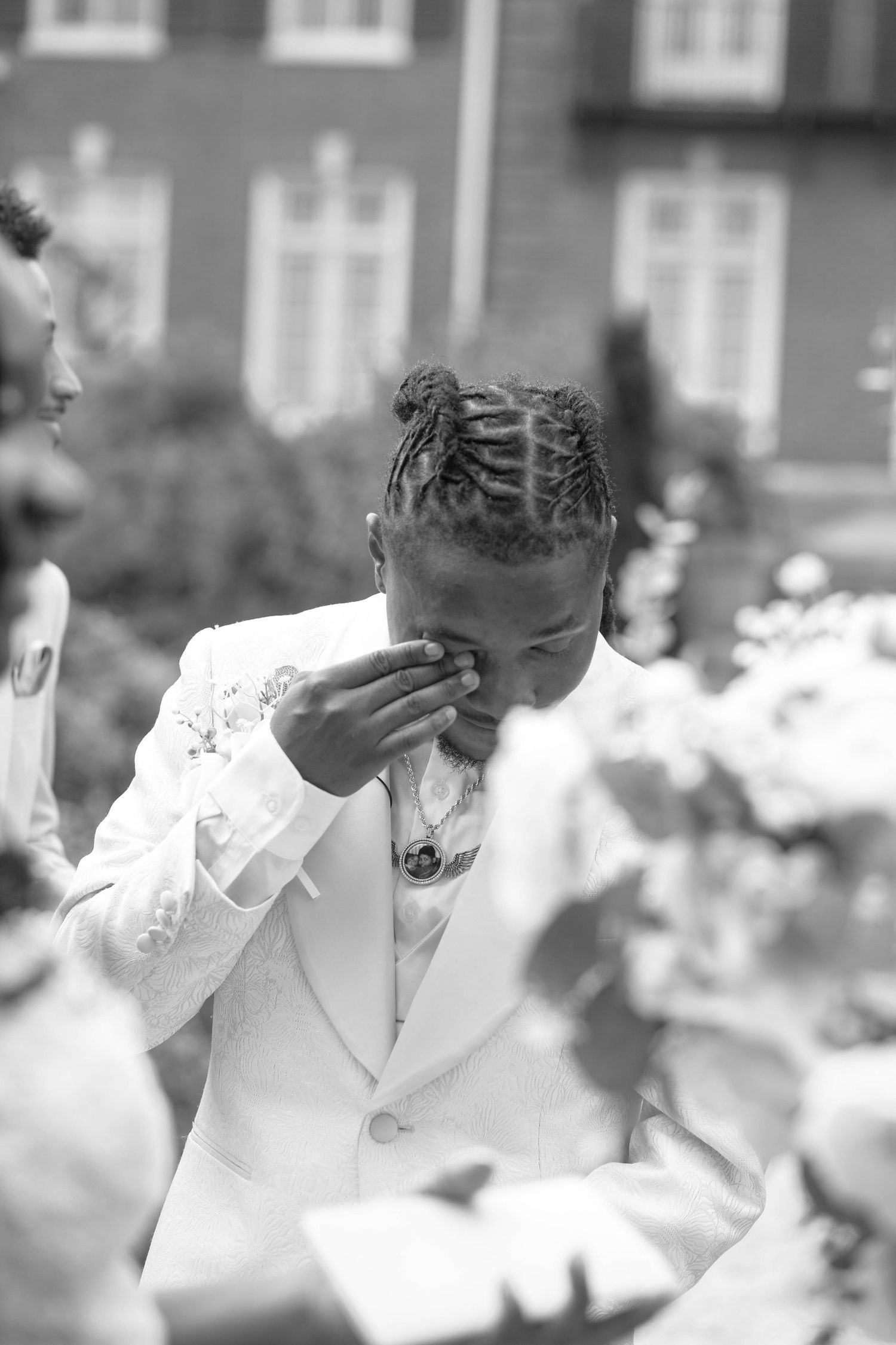 Black and white photo of emotional groom in white suit wiping tears at outdoor wedding ceremony.