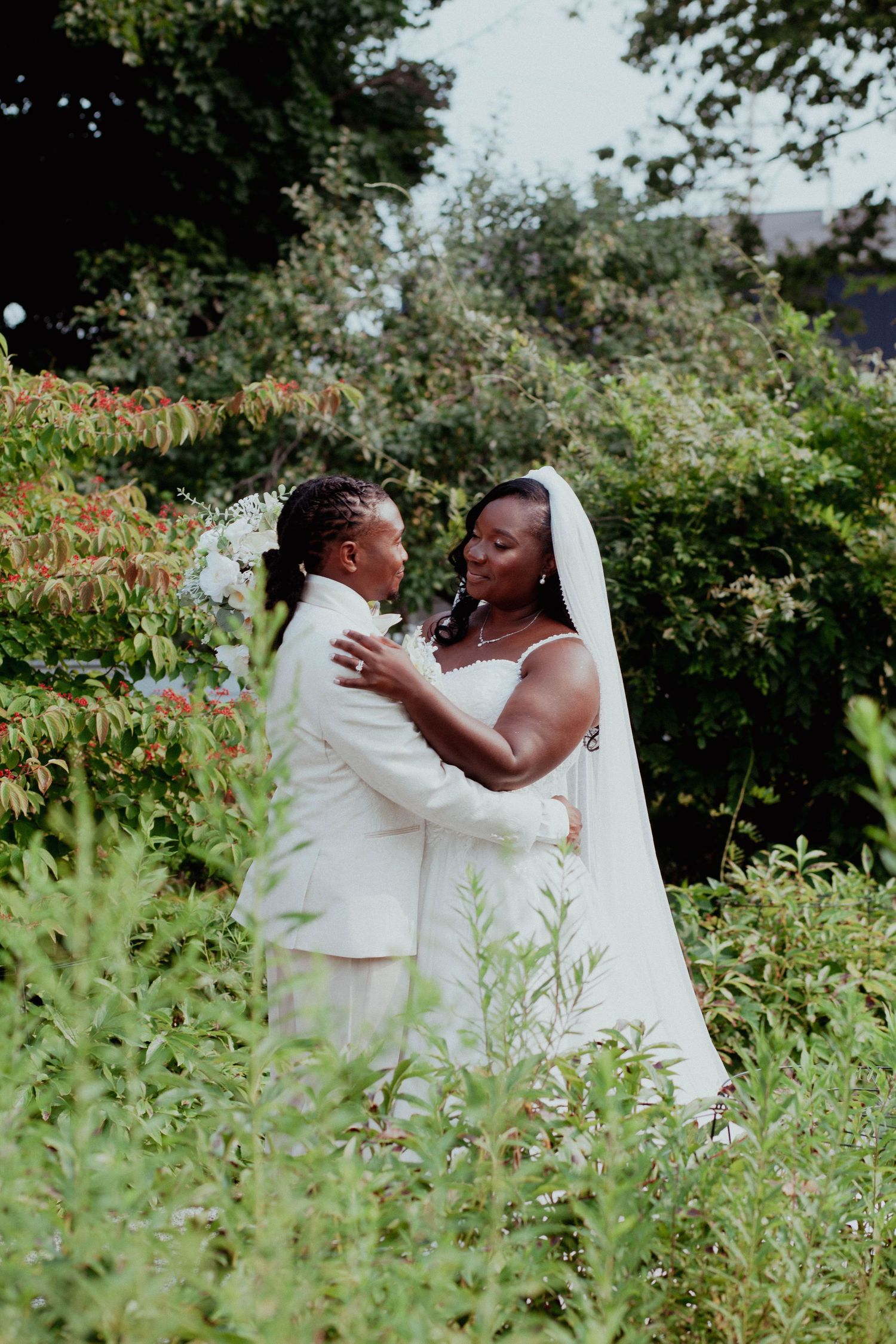 Two brides in white wedding attire embrace in a lush garden, sharing a romantic moment on their wedding day.