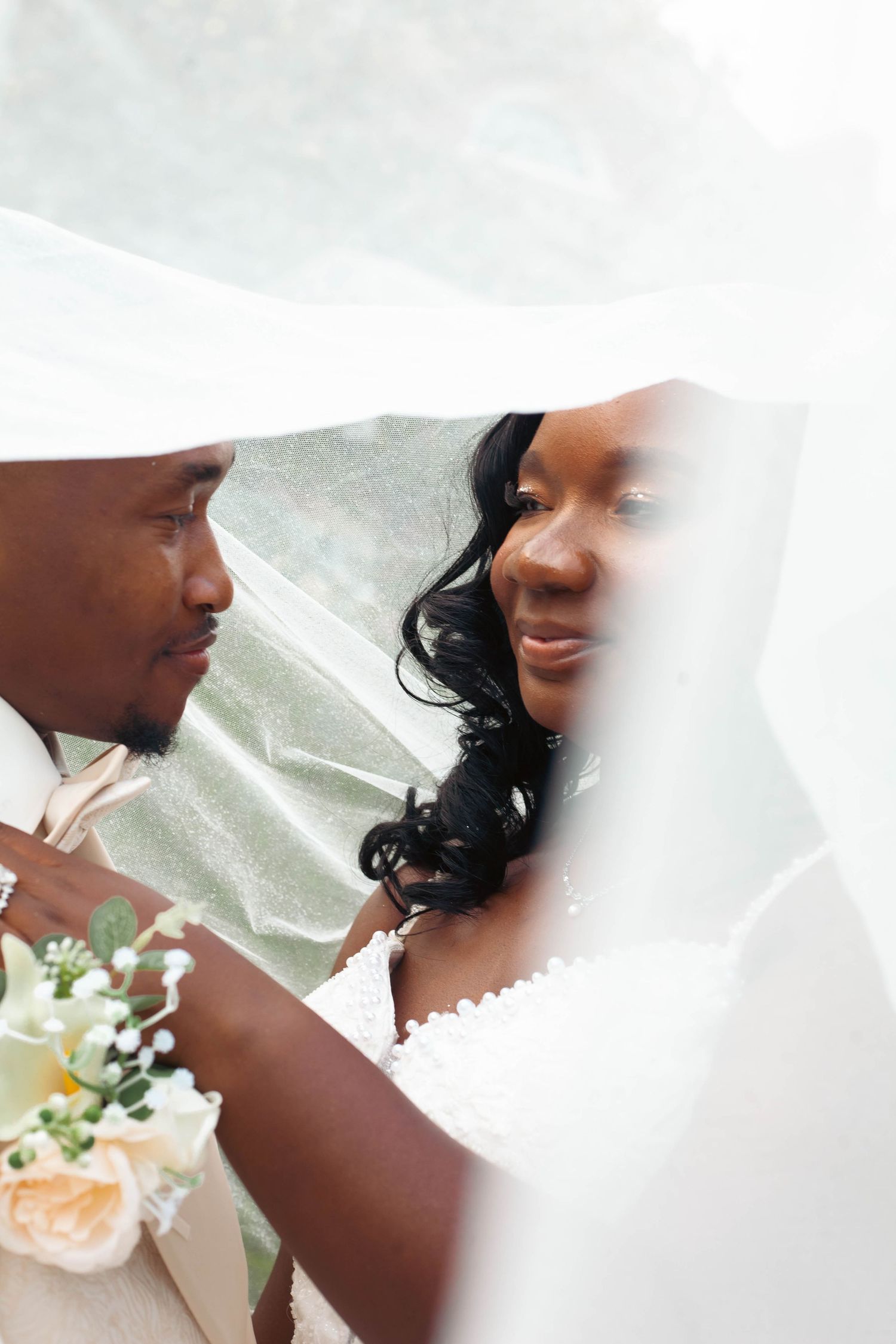 A Black bride and groom share an intimate moment under a flowing white veil, bride holding a bouquet of soft peach flowers.