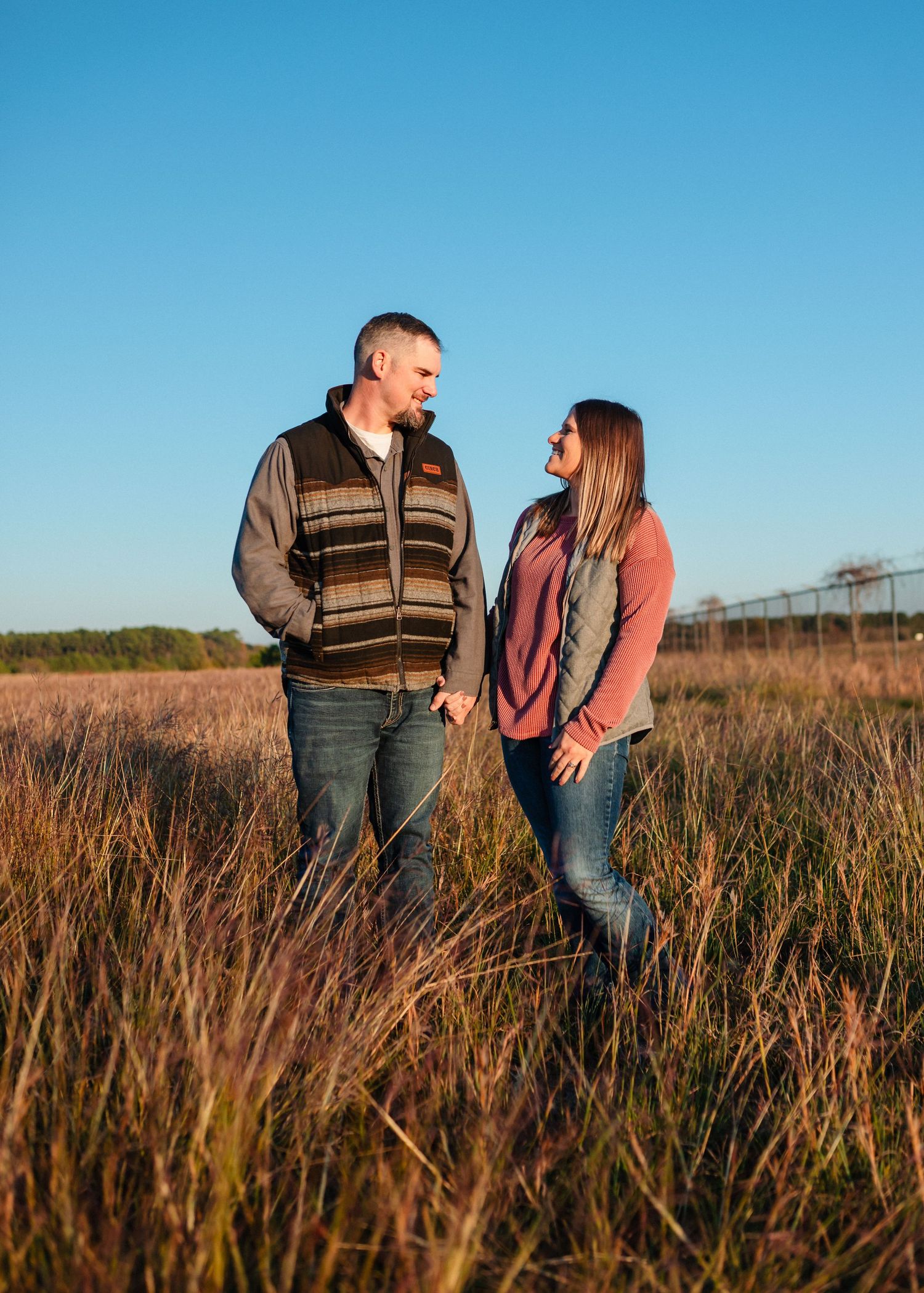 Couple standing together in tall golden grass during sunset with clear blue sky.