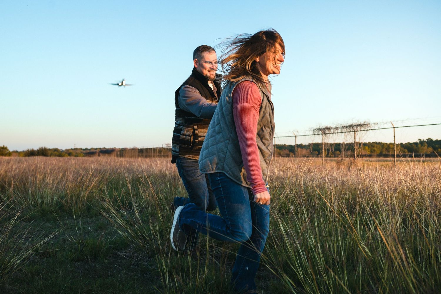 A couple embraces in a golden field at sunset with an airplane visible in the sky.