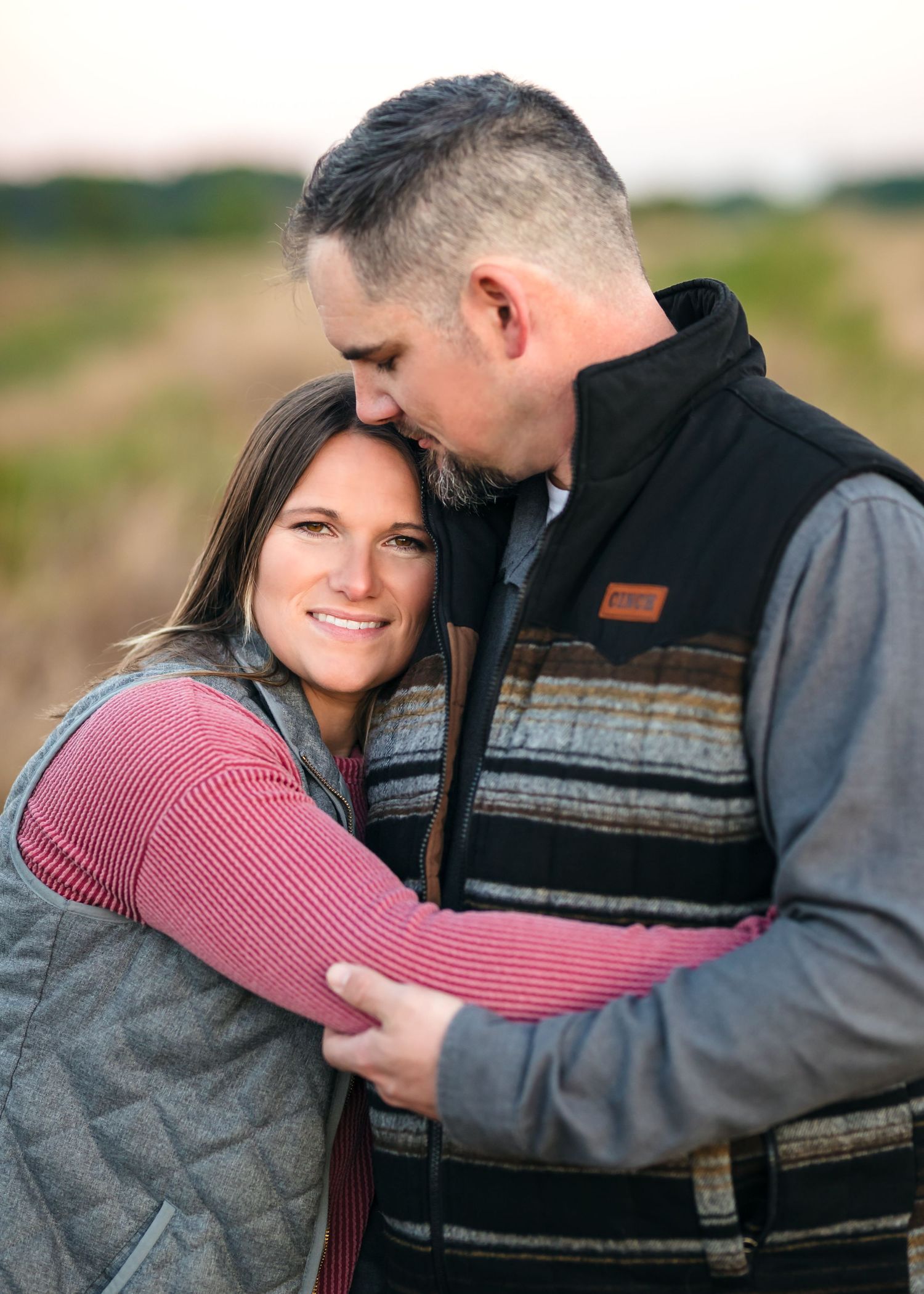 A couple embraces outdoors wearing matching quilted vests, with her in pink and him in a patterned vest.