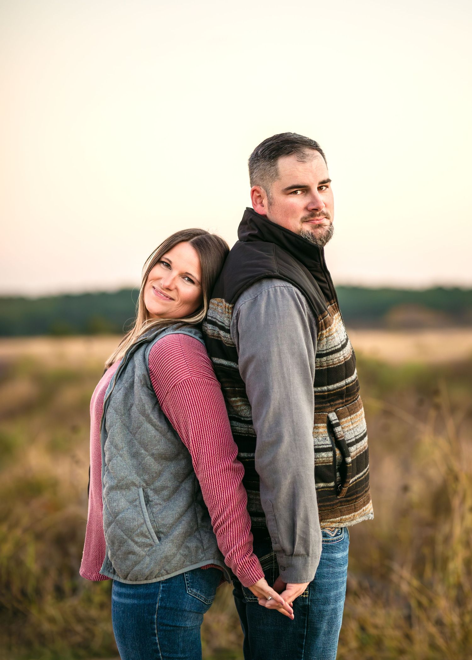 A couple shares a tender moment in a field during sunset, wearing casual attire and embracing each other.