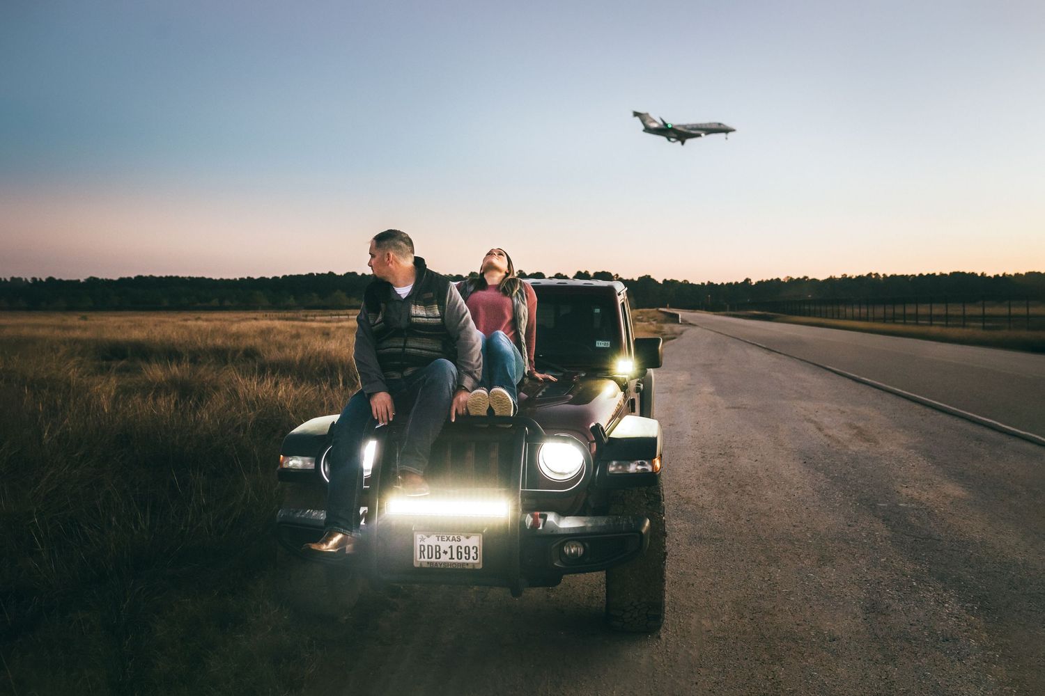 A couple relaxing on a Jeep parked near a runway with an airplane flying overhead during sunset at IAH.