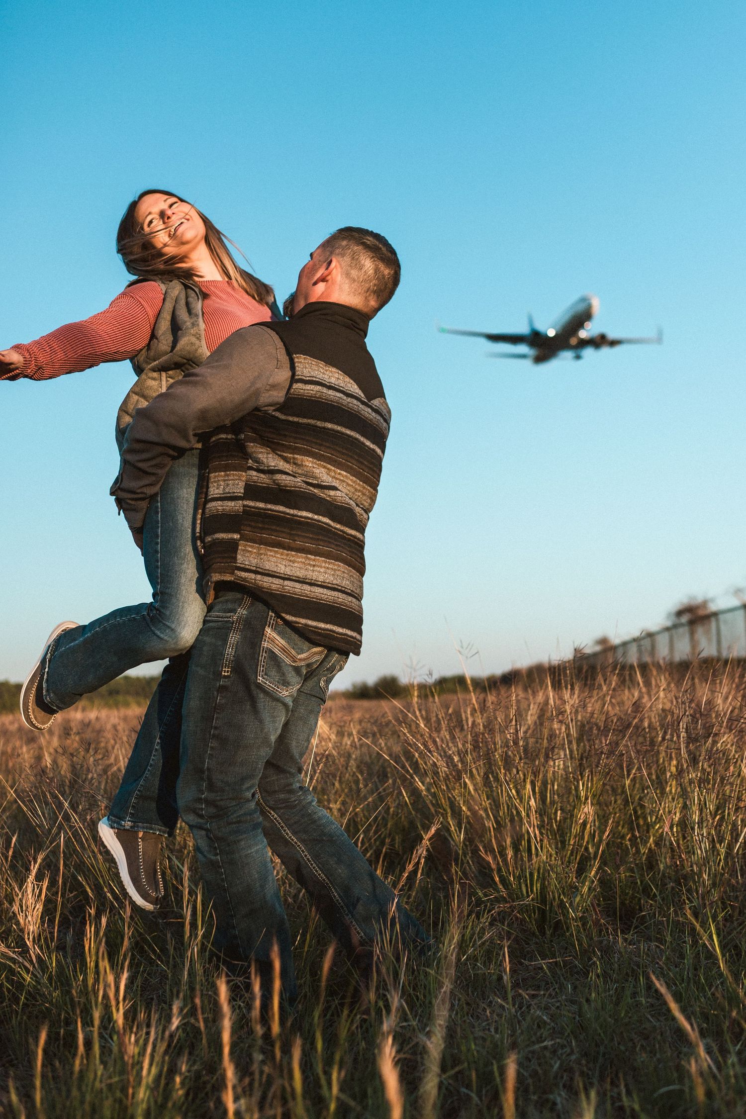 Person in brown jacket dancing in a grassy field with an airplane flying overhead in bright blue sky.