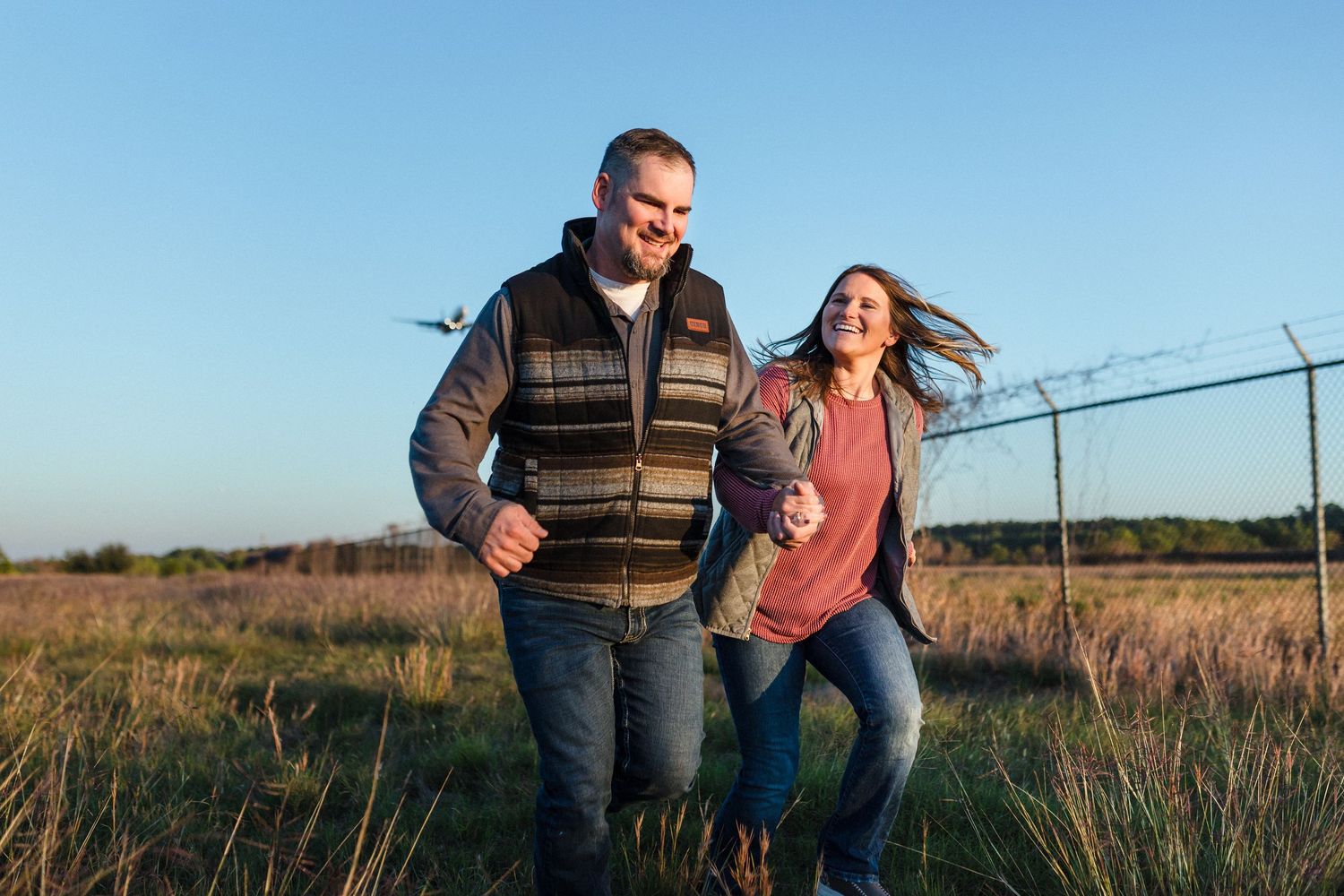 A couple runs and laughs together through a golden field at sunset, capturing a joyful candid moment.