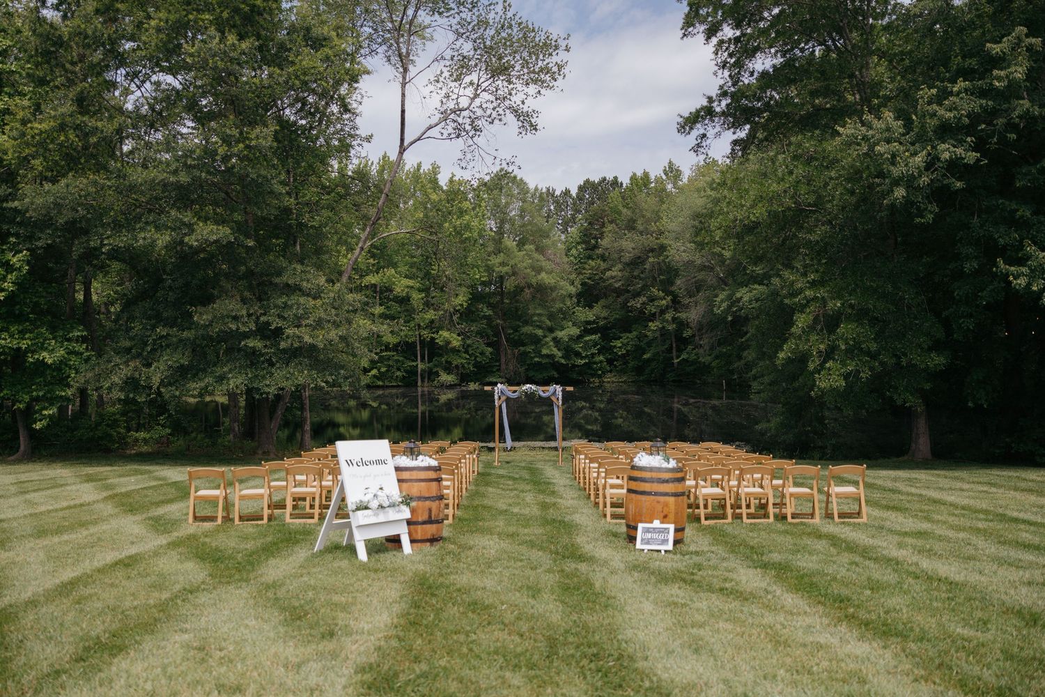 Rustic outdoor wedding ceremony setup with wooden chairs arranged on a grassy lawn surrounded by trees.