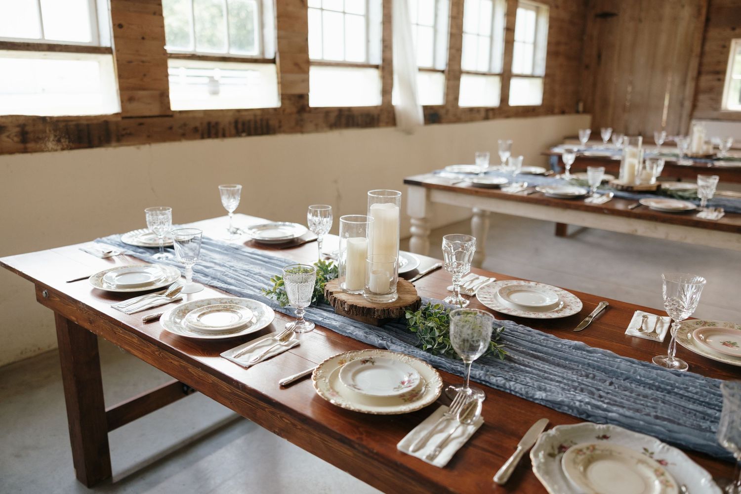 Rustic wooden dining tables set with elegant place settings in a bright, airy restaurant interior with natural lighting.