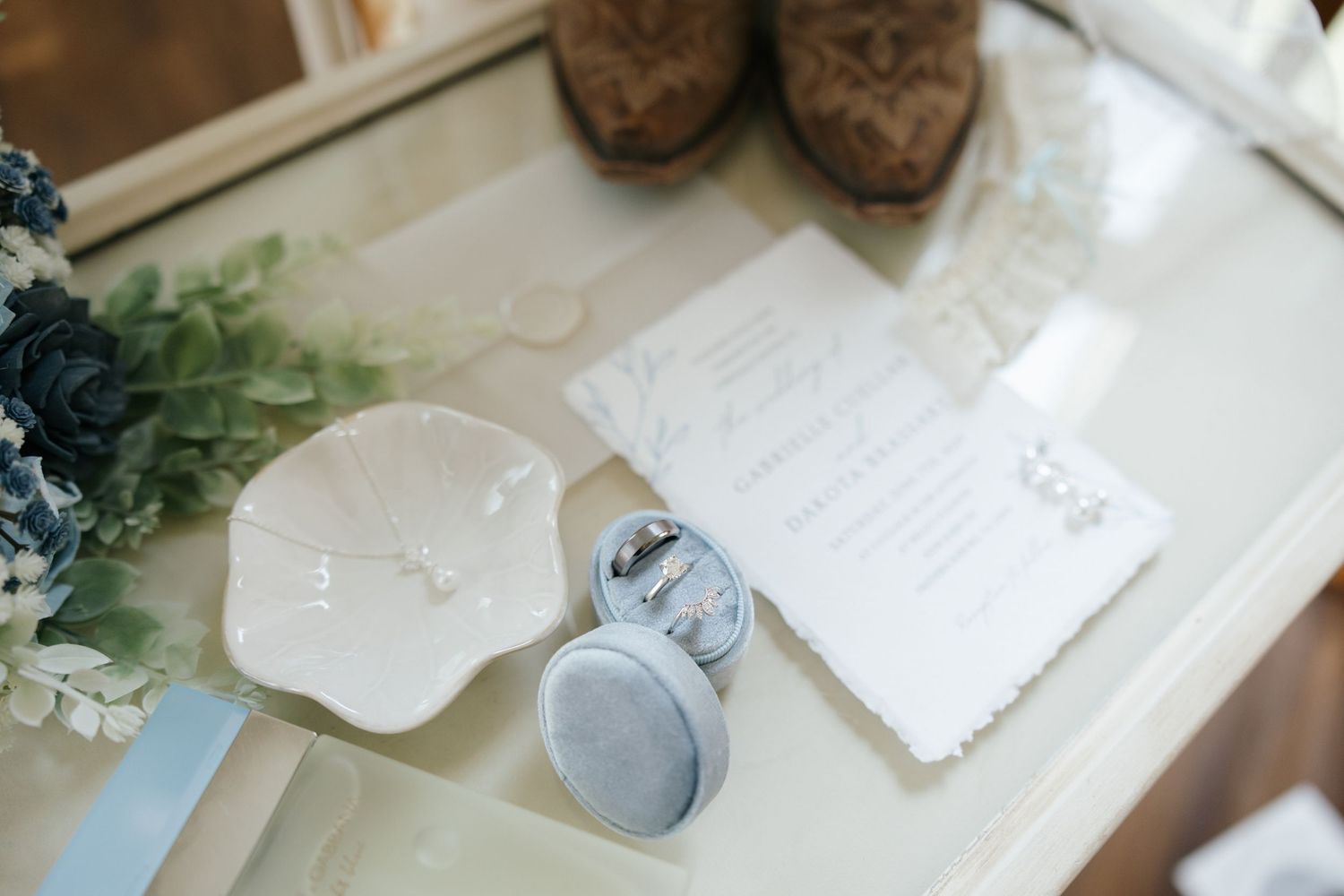Wedding details including shoes, a small white dish, and stationery arranged on a glass table with greenery decor.