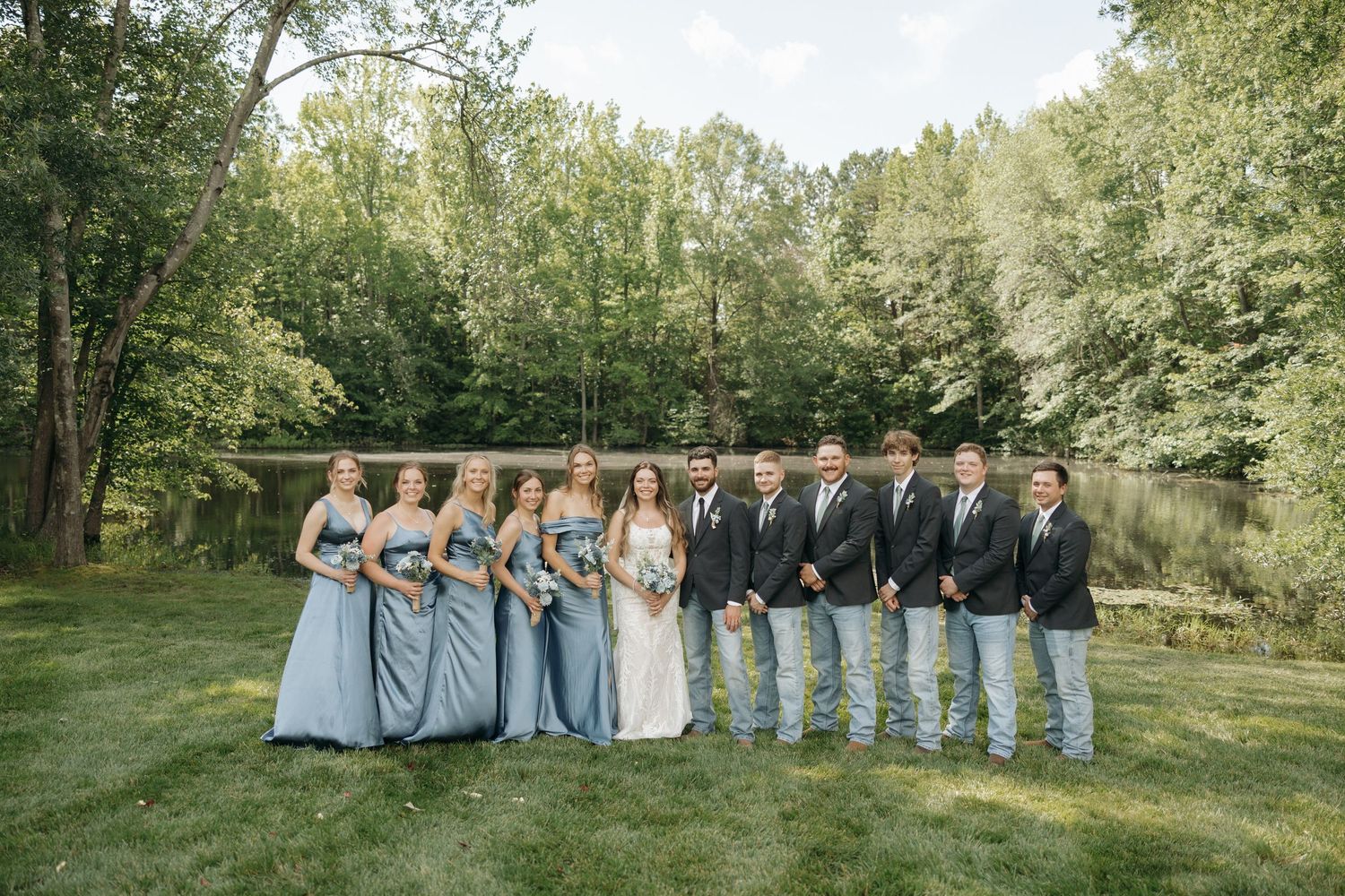 Wedding party members stand in line against a lush green outdoor backdrop during a summer celebration.