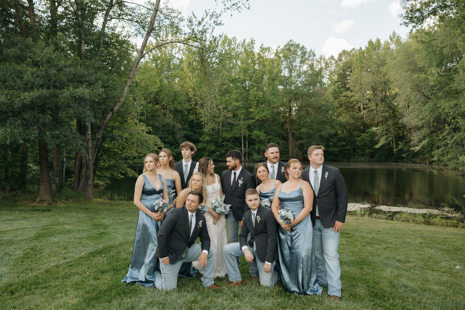 Wedding party members in light blue dresses and dark suits pose together on grassy lawn at sunset.