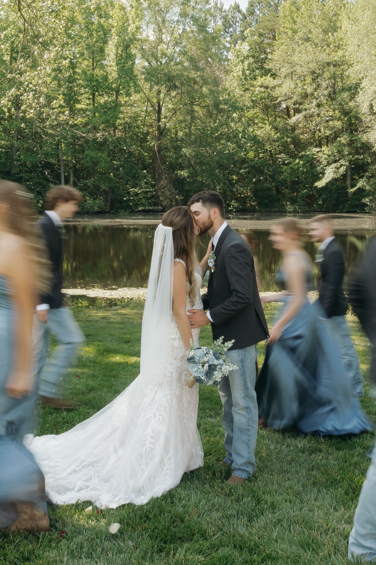 Motion blur captures a wedding party walking across a grassy lawn by a pond on a sunny afternoon.