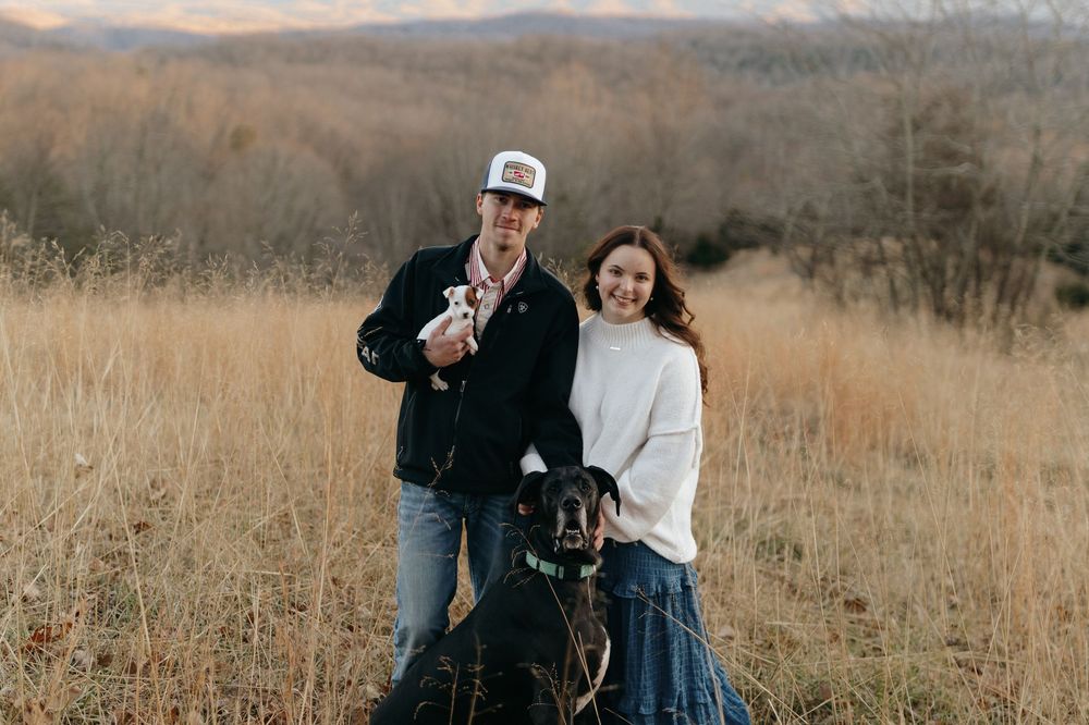 A couple and their black dog pose together in a golden field with mountains in the background during autumn.
