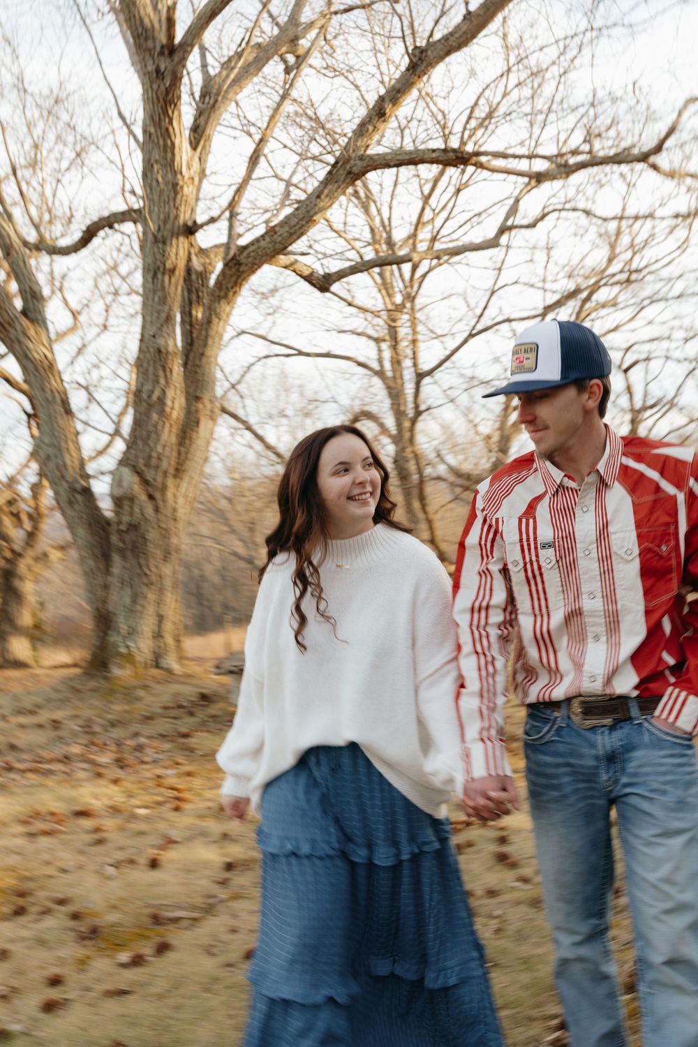 A couple shares playful moments together in a field with bare trees during an autumn photoshoot.