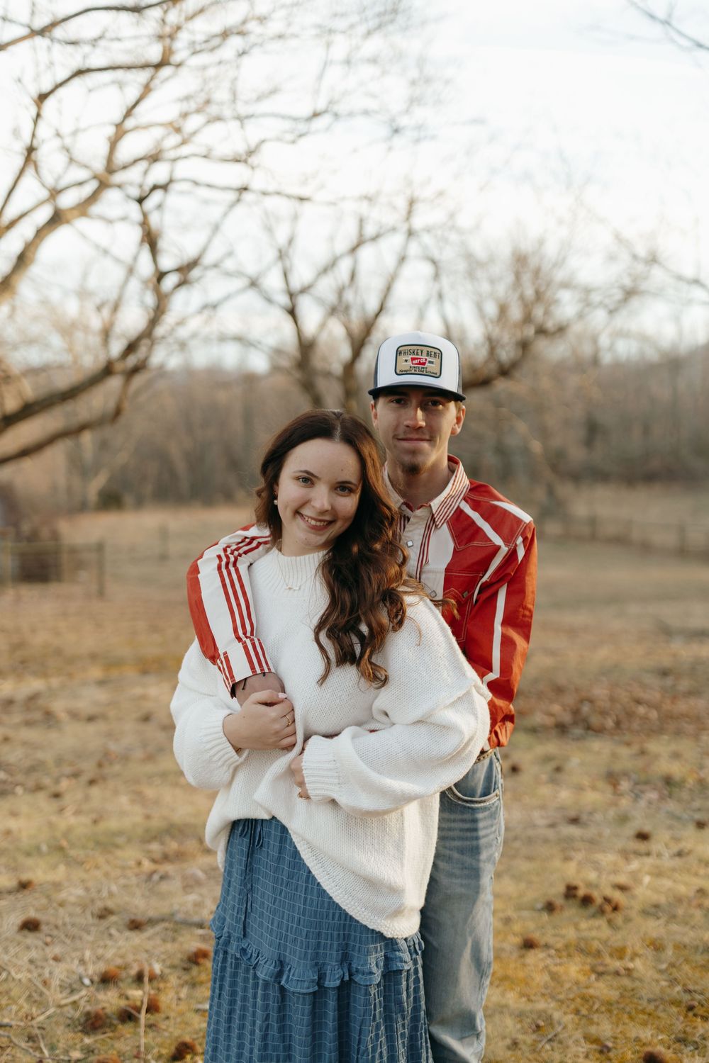 Couple in casual attire embracing in a rustic outdoor setting with bare trees in the background.