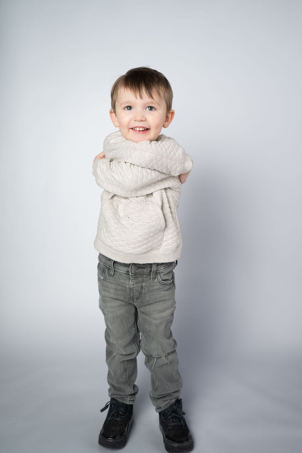 A small child wearing a white sweater and gray jeans stands against a light gray background in a playful pose.
