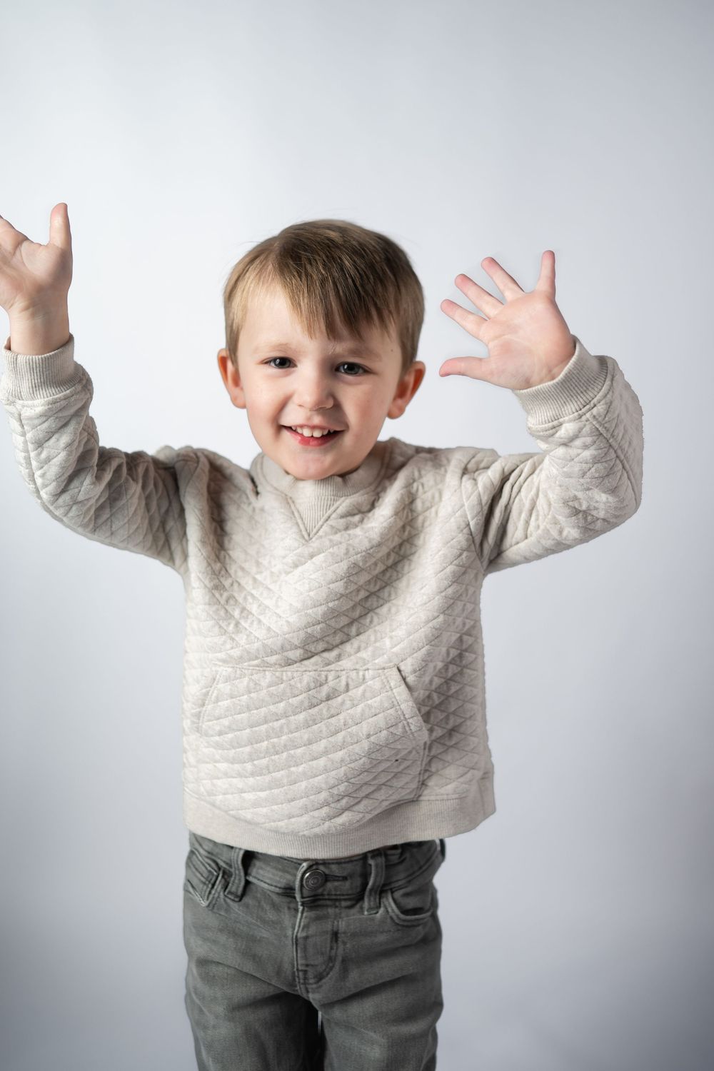 A child wearing a light gray sweater and jeans raises their arms up with excitement against a white background.
