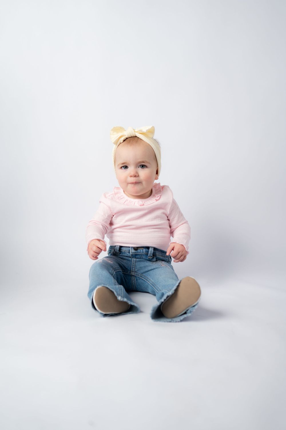 Baby wearing pink top and jeans sits on white backdrop in studio setting with natural lighting and minimalist style.