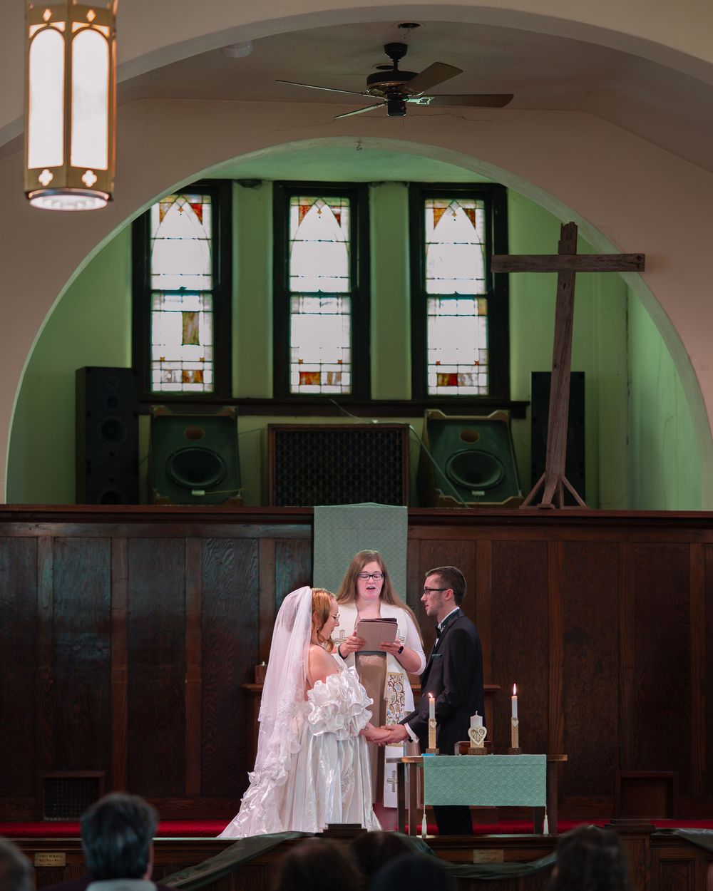 Bride and groom exchange vows at church altar with officiant, stained glass windows and wooden paneling in background.