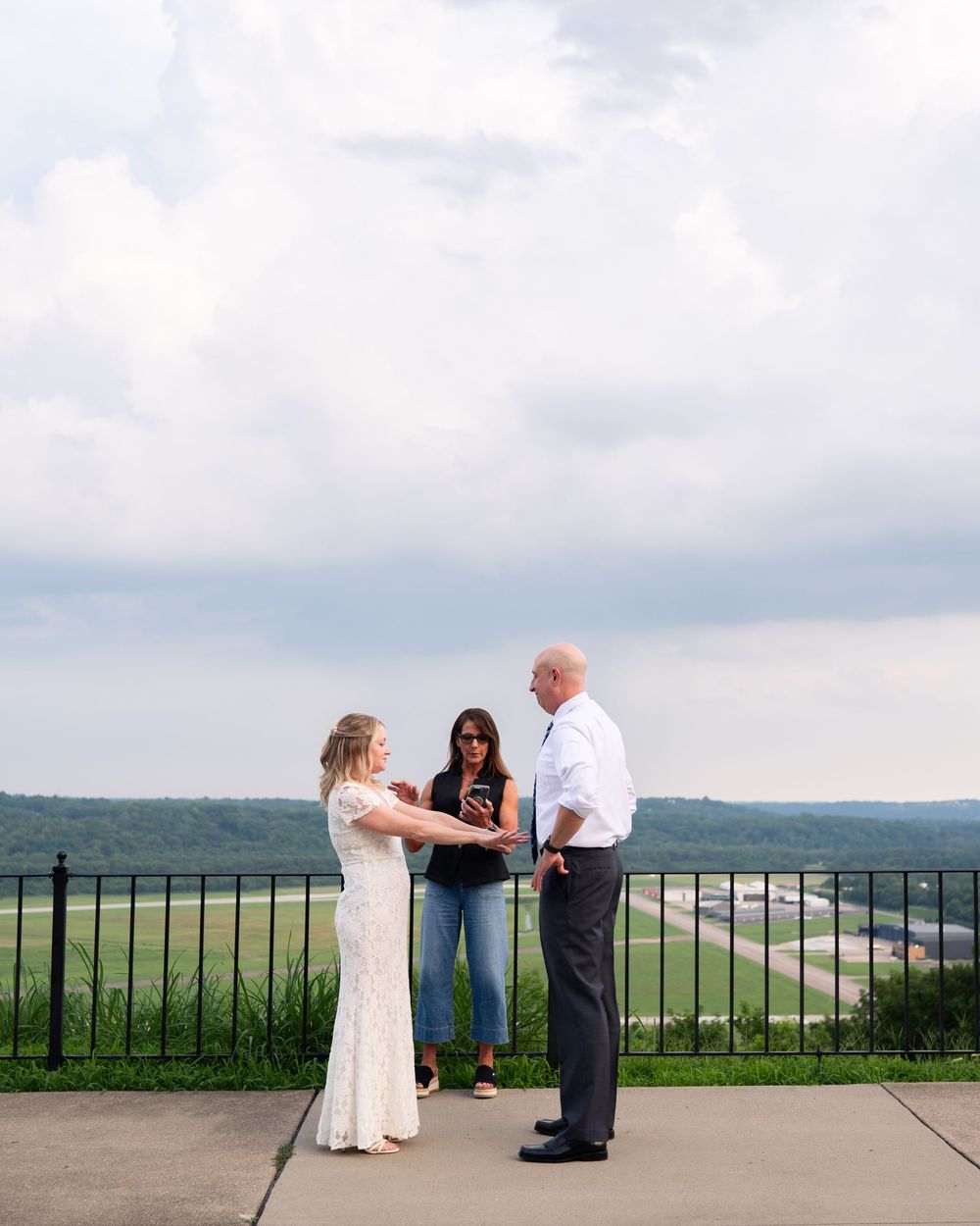 Couple exchanging vows outdoors on a scenic overlook with rolling hills, officiated by a woman in jeans.