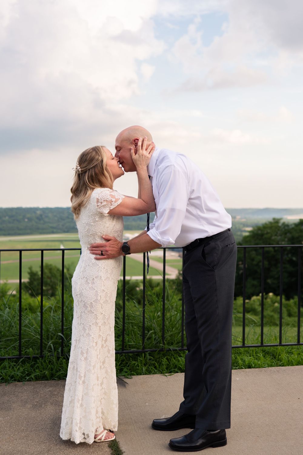 A bride in a white lace gown and groom in a white shirt share a romantic kiss on an outdoor overlook.