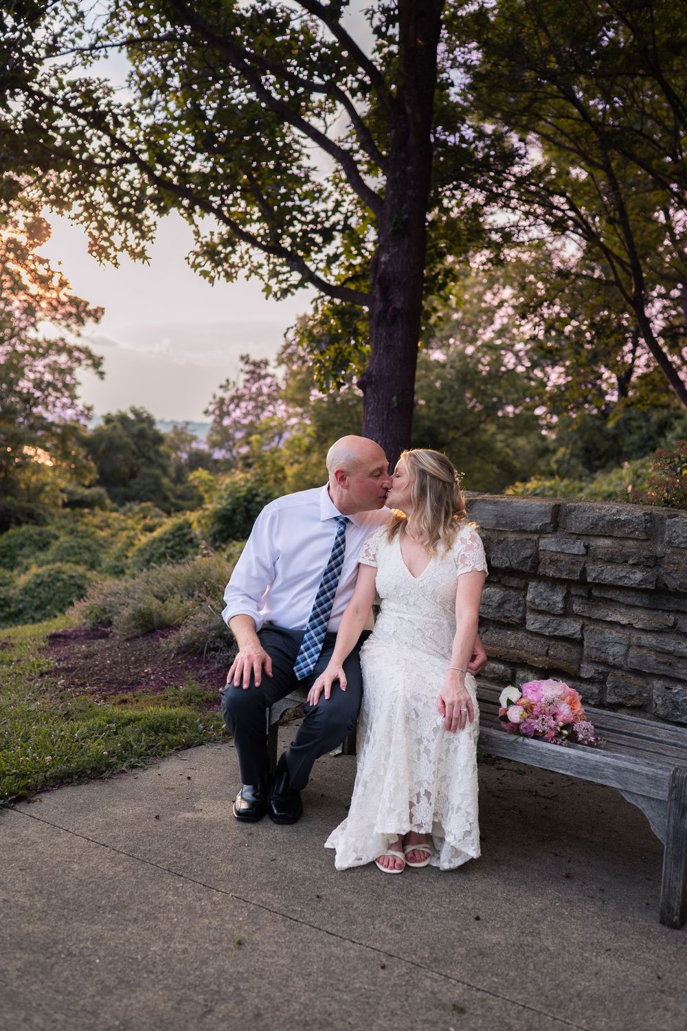 A bride and groom share a kiss on a stone bench surrounded by lush greenery at golden hour.