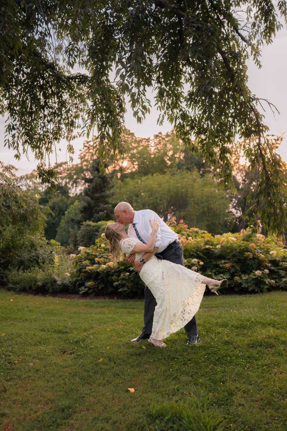 Couple sharing a romantic dip kiss in a lush garden at sunset, bride in lace gown and groom in suspenders.