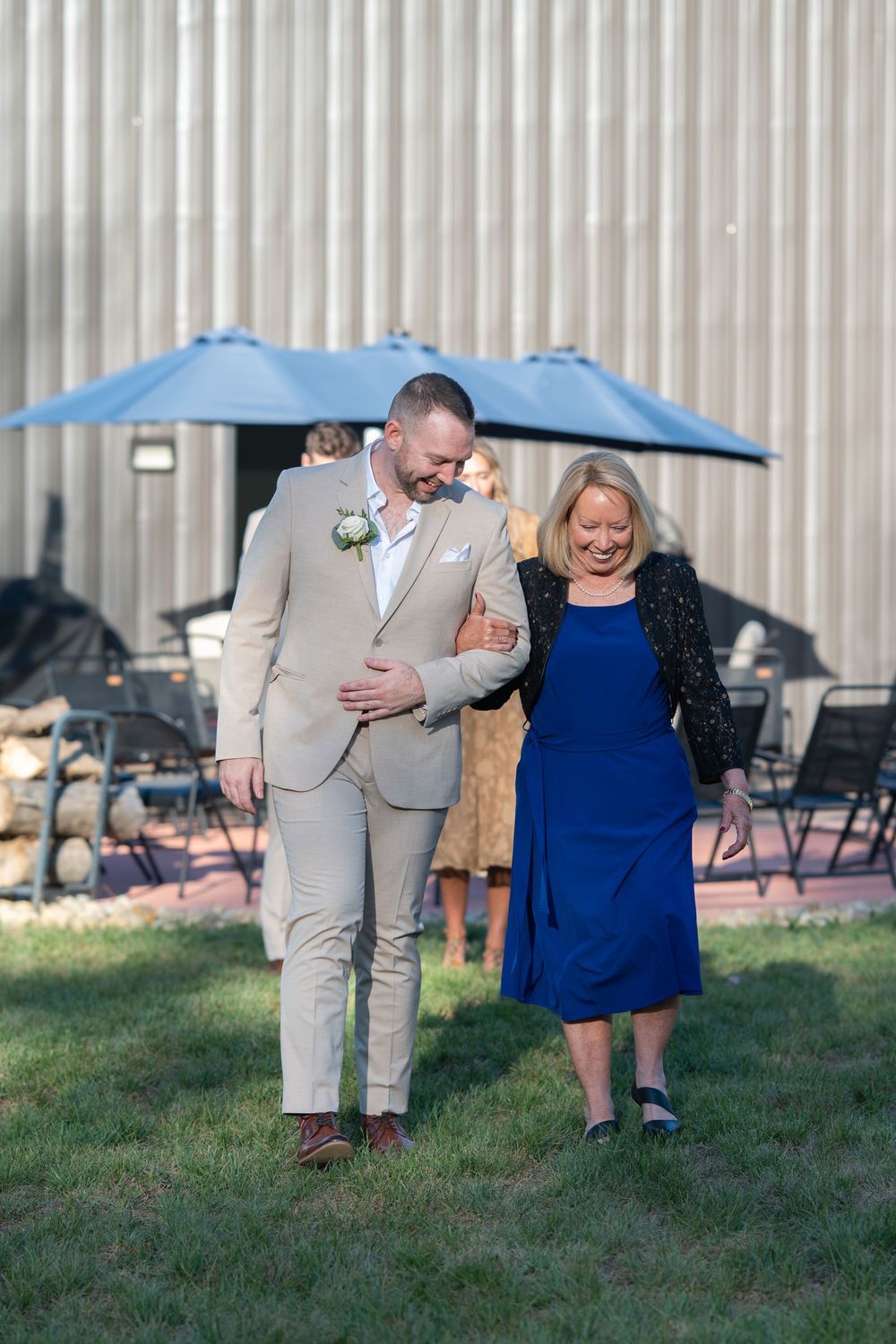 Groom in tan suit escorts smiling woman in royal blue dress outdoors at wedding venue with blue umbrellas.