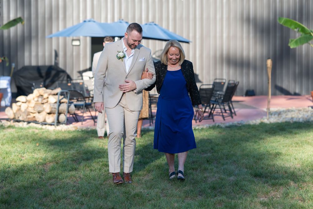 A groom in a tan suit walks with a woman in a royal blue dress outdoors at a wedding venue.