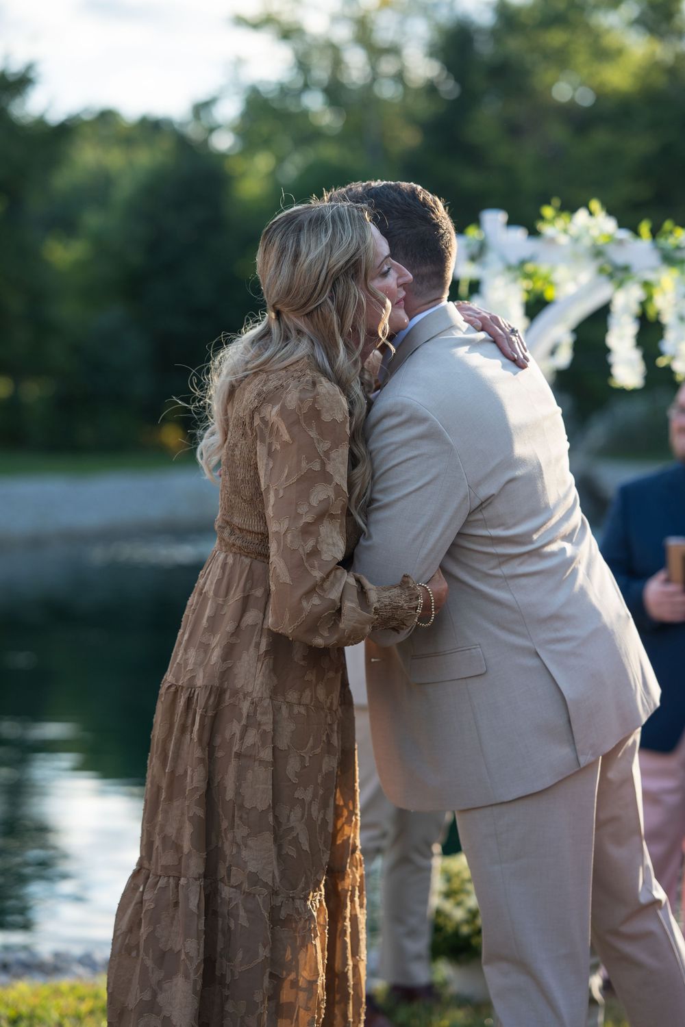 Couple sharing a romantic embrace at an outdoor wedding ceremony near a pond, bride in lace dress, groom in tan suit.