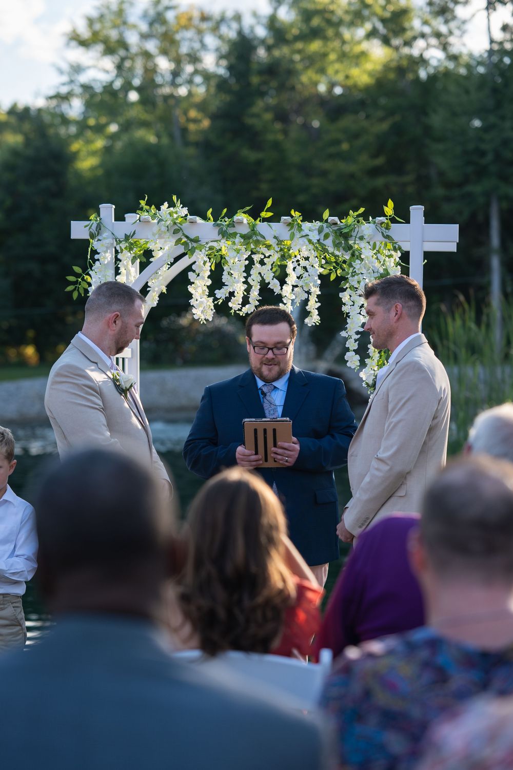Two grooms exchange vows outdoors under a floral arch with an officiant at a same-sex wedding ceremony.